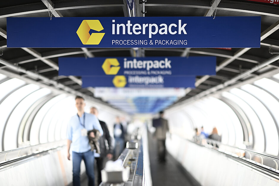 Emberek sétálnak egy mozgó járdán, amely alatt több kék tábla látható, rajtuk az interpack sárga logója és az „interpack PROCESSING & PACKAGING” felirat, egy alagútszerű szerkezetben.