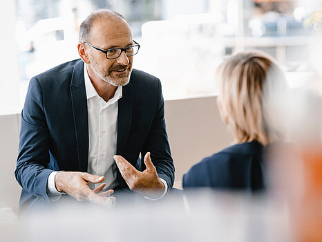 Egy sötét öltönyös, fehér ingben, szemüveges férfi, aki éppen beszélget egy nővel, aki háttal áll a kamerának.