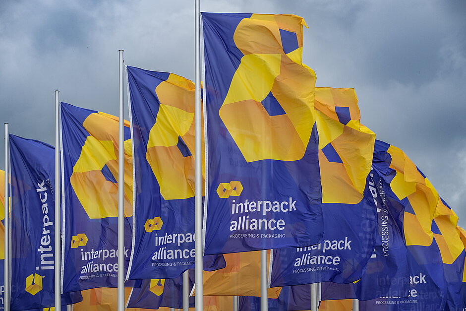 Több kék zászló lobog a szélben, sárga-narancssárga logóval és az „interpack alliance – Processing & Packaging” felirattal, felhős égbolton.