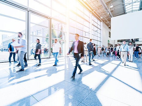 Egy világos, napfényes kiállítóterem, ahol sokan sétálgatnak a tágas térben, miközben a nagy ablakok árnyékokat vetnek a padlóra.